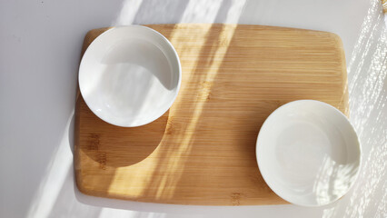 Cutting board made of natural bamboo and white plate saucer on table with shade and light from sun. Abstract natural background, texture, frame, copy space. Concept of cooking with beautiful things