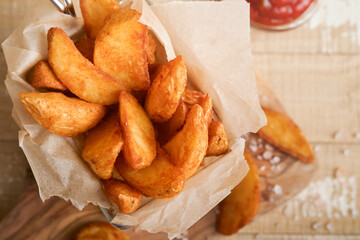French fries in metal wire basket with salt and ketchup on old wooden light background clous up. Fried potatoes. Fast food and unhealthy food concept.