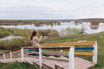 A young woman stands on a wooden path in a spring park, spending time outdoors, drinking coffee or tea. A pretty woman in a shirt is relaxing in nature. Recreation and leisure concept.