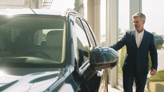 Male driver opening door while luk the interior of his nev auto. Serious middle age customer standing near new car in auto show room, looking at car interior and examining carefully vehicle
