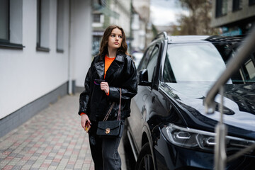 Obraz na płótnie Canvas Young girl in leather jacket posed in urban city, holding credit card with handbag and phone against black business car.