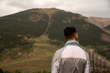 Naklejka premium A young dark-haired man with his back turned and wearing a grey sweatshirt admiring the mountainous and natural landscape of Huesca in Aragon on a cloudy day.