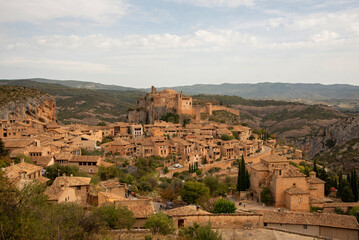 Obraz premium Vista panorámica del turístico pueblo medieval de Alquezar lleno de pequeñas casas de ladrillo y una gran iglesia en la parte superior rodeada por un paisaje montañoso en un día claro.
