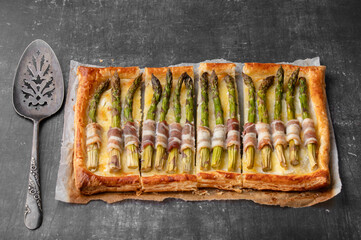 Puff pastry tart with green asparagus and bacon. Asparagus pie with cheese and bacon on a dark background