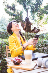 Woman laughing loudly with a mobile phone in her hand sitting on a terrace in a park