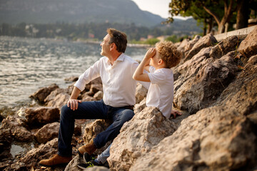 Fototapeta na wymiar Brunette father sitting with his blondie curly hair son at the stones of Italian Garda lake looking alongside 
