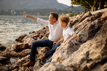 Brunette father pointing with finger sitting with his blondie curly hair son at the stones of Italian Garda lake 