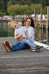 Brunette mom and blondie curly hair son having fun and smiling widely at the wooden pier in Italian Garda lake port 