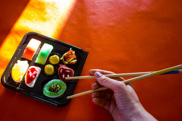 Sushi sticks in a woman's hand against a sushi candy set on a red background view from above. Oriental food. Exotic dessert, sweets. Asian food eating process. Japanese rolls with fish, seafood.
