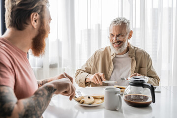 Cheerful gay man talking to blurred tattooed partner during breakfast at home.