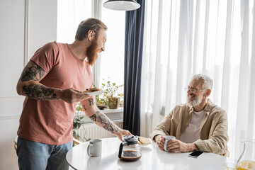 Smiling gay man looking at tattooed partner holding pancakes at home. 