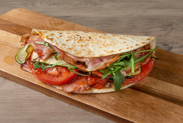 Traditional Italian piadina with pork, porchetta tomato zucchini arugula photographed up close above a cutting board at a rustic wooden table. horizontal close up