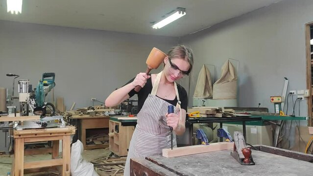 Craftswoman at Work: A focused young woman, wearing protective eyewear and an apron, meticulously carves wood with a chisel and mallet in a workshop, surrounded by tools and materials.