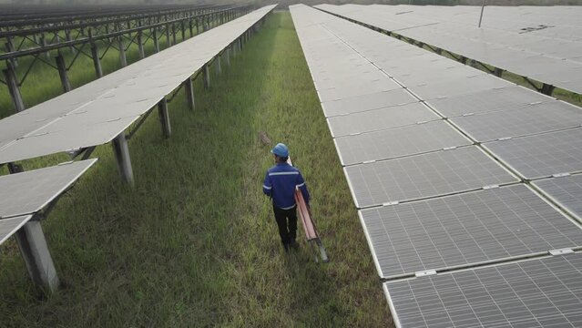 Solar Farm Worker Inspecting Panels: A solitary worker in a blue uniform walks between rows of photovoltaic solar panels in a vast solar farm.