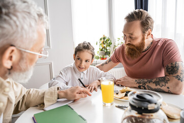 Preteen girl writing on notebook near homosexual parents and pancakes at home. 