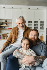 Homosexual man hugging smiling daughter near partner and looking at camera at home. 