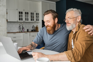 Smiling homosexual man using laptop and hugging partner with credit card at home. 