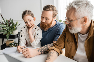 Preteen child writing on notebook near tattooed same sex parents at home. 