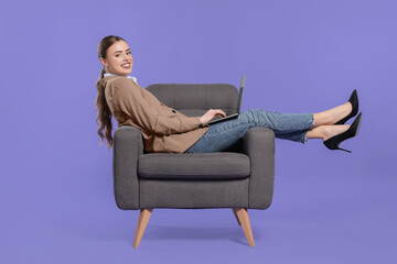 Fototapeta na wymiar Happy woman with laptop sitting in armchair on violet background
