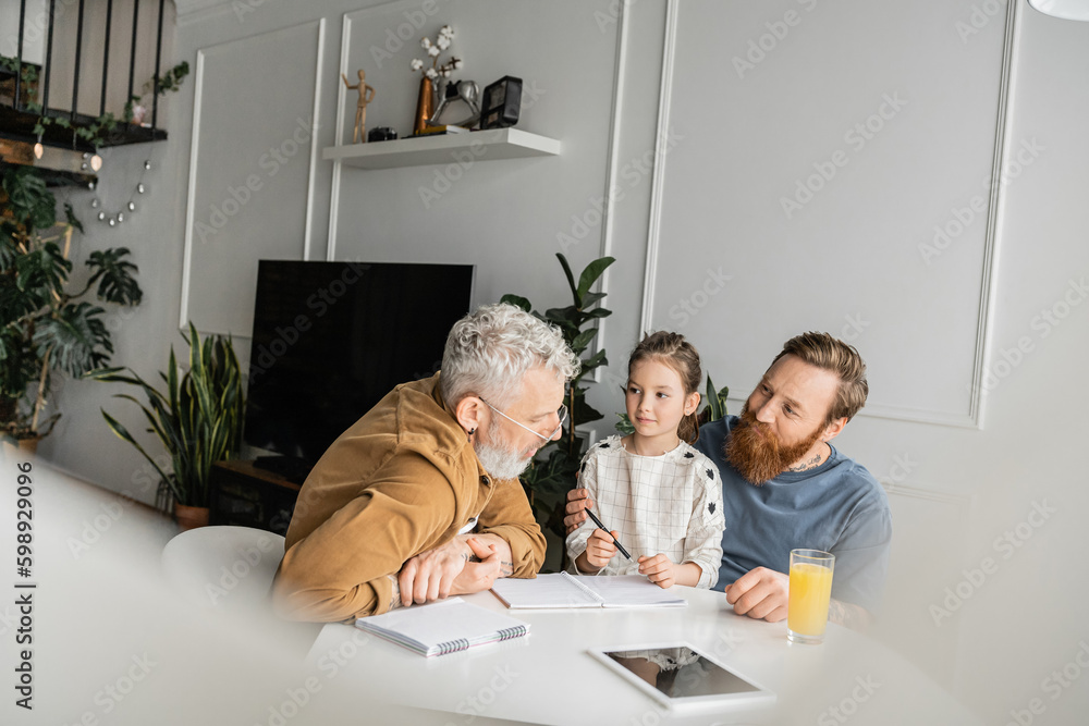 Wall mural Gay man helping daughter with homework near partner and digital tablet at home. 