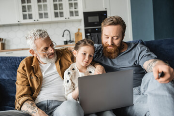 Preteen child using laptop near tattooed gay fathers at home. 