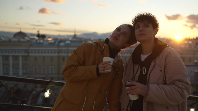 4k Two lgbt women drink coffee on rooftop in city spbd. Young lesbian couple drinking hot drink and looking with smiles, hugging and standing on background of town during sunset. Happy non-binary