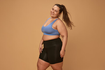 Obraz premium Portrait of young, smiling, overweight woman posing in comfortable sportswear, leggings and top against brown studio background. Concept of sport, body-positivity, weight loss, body and health care