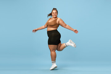Young, happy, active, overweight woman training in sportswear against blue studio background. Self-love and acceptance. Concept of sport, body-positivity, weight loss, body and health care