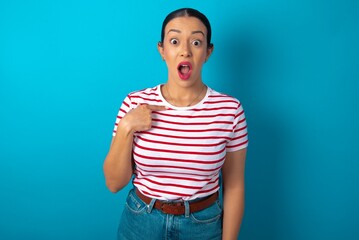 beautiful woman wearing striped T-shirt over blue studio background being in stupor shocked, has astonished expression pointing at oneself with finger saying: Who me?