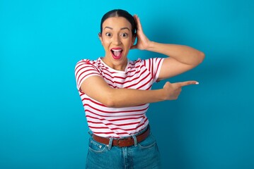 Naklejka premium beautiful woman wearing striped T-shirt over blue studio background feeling positive has amazed expression, indicates something. One hand on head and pointing with other hand.
