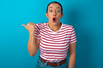 Shocked beautiful woman wearing striped T-shirt over blue studio background points with thumb away, indicates something. Check this out. Advertisement concept.