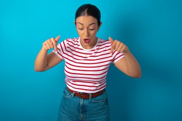 Naklejka premium Amazed beautiful woman wearing striped T-shirt over blue studio background points down with fore fingers, opens mouth being shocked. Advertisement concept.