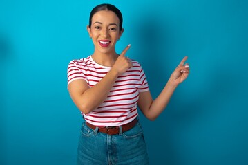 Positive beautiful woman wearing striped T-shirt over blue studio background with beaming smile pointing with two fingers and looking on empty copy space. Advertisement concept.