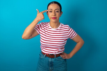 Naklejka premium Unhappy beautiful woman wearing striped T-shirt over blue studio background curves lips and makes suicide gesture, shoots in temple with hand, tries kill himself.