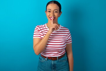 Smiling beautiful woman wearing striped T-shirt over blue studio background makes shush gesture,...