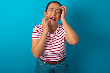 Young gloomy beautiful woman wearing striped T-shirt over blue studio background, hiding face with hands pouting and crying, standing upset and depressed complaining about job problem.