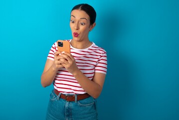 beautiful woman wearing striped T-shirt over blue studio background looks with bugged eyes, holds modern smart phone, receives unexpected message from friend, reads reminder.