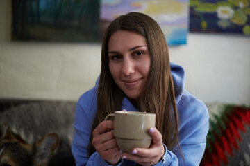 Friendly young Ukrainian woman sitting on a couch with a cup of coffee. Portrait of a pretty white brunette female drinking hot beverage in a cafe