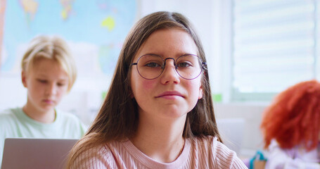 Portrait of pretty Caucasian teen clever schoolgirl in glasses sitting at desk in classroom and looking at camera at lesson. Among kids. Indoor. Little girl learning in school concept. Close up.