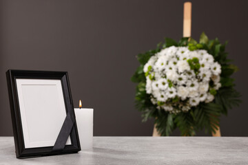Photo frame with black ribbon, burning candle on light table and wreath of flowers near grey wall...