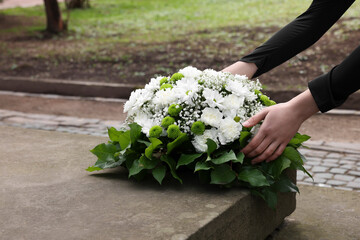 Woman with wreath of flowers near tombstone outdoors, closeup. Funeral attribute