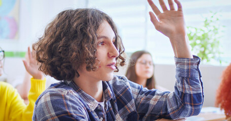 Close up of face of Caucasian teen clever schoolboy with long hair sitting in classroom and rising hand to answer at lesson. Indoor in class. Little smart boy learning in school concept.