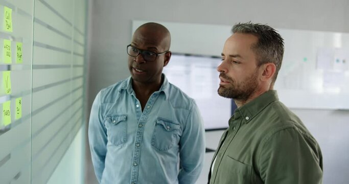 Two diverse businessmen smiling during a brainstorming session together with adhesive notes on a glass wall of an office