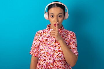 beautiful woman wearing floral dress over blue studio background making hush gesture with finger on her lips wearing  wireless headphones. Be quiet.