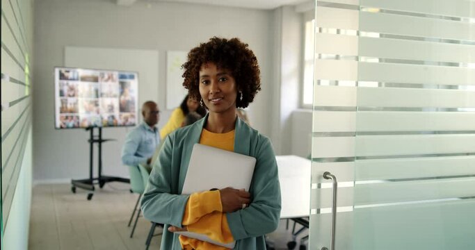 Smiling young businesswoman standing with a laptop at the door to a boardroom with coworkers meeting in the background