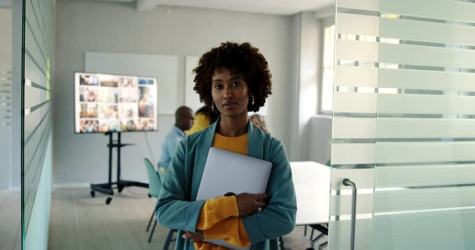 Confident young businesswoman standing with a laptop at the door to a boardroom with coworkers meeting in the background