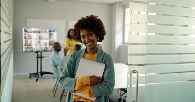 Laughing young businesswoman standing with a laptop at the door to a boardroom with coworkers meeting in the background