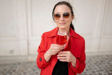 Frau, 40+, mit Sonnenbrille im roten Mantel mit einem Glas Spritz