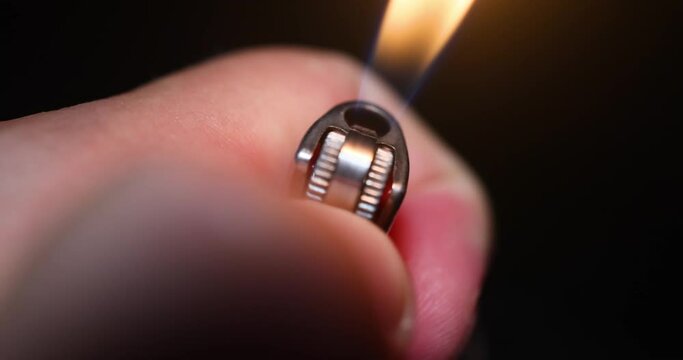 A man's hand holds a cigarette lighter, igniting a flame that illuminates the darkness around it. The flickering flame casts dancing shadows as it provides light, while the man's hand carefully contro