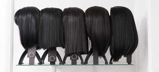 Showcase of natural looking wigs in different haircut options fixed on the wig holders in beauty salon. Row of mannequin heads with black asian hair on shelf in wig shop
